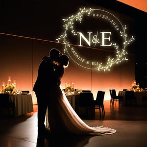 May include: A bride and groom kiss in front of a wall with a wedding monogram. The monogram is a circle with a floral wreath and the letters N&E, with the text "Natalie & Eli" and the date "04/20/2024".