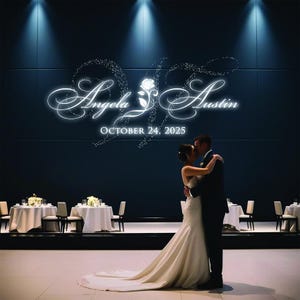 May include: A couple embraces on a dance floor in front of a wall with the names "Angela & Austin" and the date "October 24, 2025" illuminated in white. The bride wears a white wedding dress.