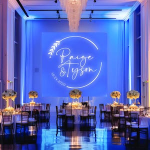 May include: A wedding reception venue with white floral centerpieces on tables with chairs. A large neon sign on the wall reads "Paige & Tyson 10.24.2020".