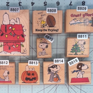 May include: A collection of eight wooden rubber stamps featuring Peanuts characters, including Snoopy and Woodstock, with labels 8807 through 8815. The stamps are arranged on a cutting mat with inch markings, displaying various scenes like a doghouse, pumpkin, and sports.