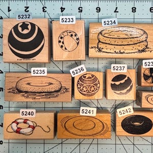 May include: A collection of wooden rubber stamps featuring beach and summer-themed designs. The stamps include images of a beach ball, a swimming pool, a life preserver, and other related objects. The stamps are mounted on wooden blocks.