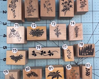 May include: A collection of wooden stamps with botanical designs. The stamps feature various plant illustrations, including leaves, branches, and floral patterns. The stamps are arranged on a blue and green cutting mat.
