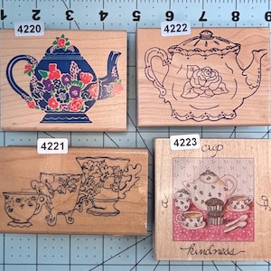 May include: Four wooden stamps with tea-themed designs. One stamp features a blue teapot with floral accents. Another shows a teapot outline with a rose. The third depicts teacups and a vase. The last stamp has a teapot, cups, and the word "kindness".