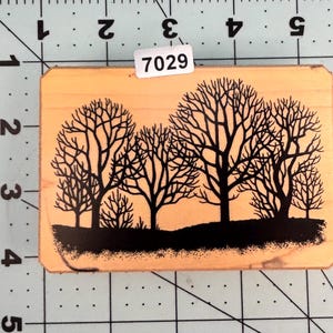 May include: A rectangular wooden stamp featuring a black silhouette of several bare trees against a black horizon line. The stamp has the number "7029" on a white sticker. The stamp is made of light-colored wood.
