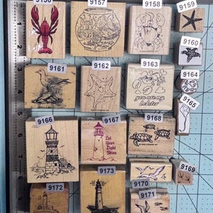 May include: A collection of wooden rubber stamps featuring nautical and marine themes, including lighthouses, seagulls, crabs, lobsters, sea turtles, and starfish, arranged on a cutting mat with a metal ruler showing measurements in inches.