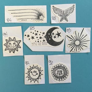 May include: A set of ten black and white iron-on patches with various designs, including a shooting star, a winged star, a crescent moon with stars, a sun with a face, and a sun with rays. The patches are on white cardstock with handwritten numbers on each.