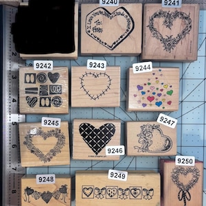 May include: A collection of eleven wooden rubber stamps featuring various heart-themed designs, including floral wreaths, stitched patterns, and decorative borders. Each stamp is labeled with a number from 9240 to 9250, arranged on a grid mat.