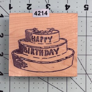 May include: A wooden stamp featuring a two-tiered birthday cake design. The words "HAPPY BIRTHDAY" are printed across the cake layers. The stamp also includes the text "TO:" on the bottom layer. A small sticker with the number "4214" is on the top.