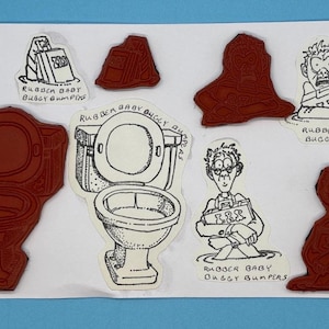 May include: A set of nine red rubber stamps with black ink illustrations of a toilet, a person holding a book, a person with a book, a person with a book, a person with a book, a person with a book, a person with a book, a person with a book, and a person with a book. The stamps are labeled "Rubber Baby Buggy Bumpers".