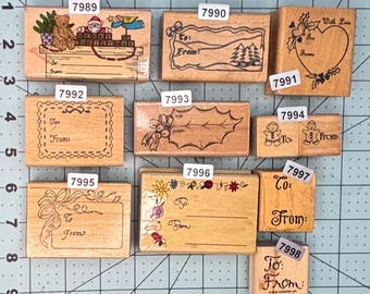 May include: A collection of ten rectangular wooden rubber stamps for gift tags, each labeled with a number from 7989 to 7998. The stamps feature various holiday designs including teddy bears, holly, gingerbread people, and floral borders with To and From lines.