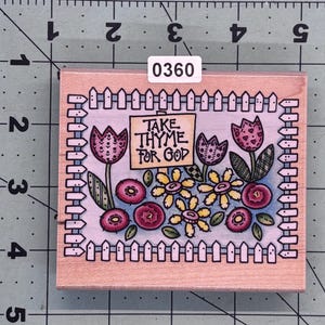 May include: A wooden stamp with a colorful illustration of a garden scene. The stamp features a sign that reads "Take Thyme For God" with flowers and a picket fence.
