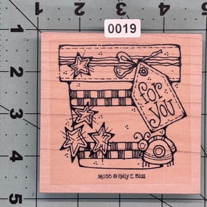 May include: A pink wooden rubber stamp with a black and white illustration of a gift box with stars, stripes, and a tag that says "for you". The stamp is labeled "M058 Holly E. Bliss".
