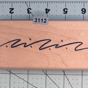May include: A rectangular wooden stamp with a black wavy line design. The stamp has the number "3112" on a small sticker. The stamp is on a gray and white grid background.