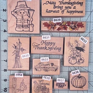 May include: A collection of eleven wooden rubber stamps featuring Thanksgiving themes, including a pilgrim, pumpkins, corn, acorns, and text such as 'Happy Thanksgiving' and 'Happy Turkey Day', arranged on a cutting mat with inch markings.