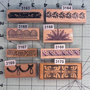 May include: A collection of eight wooden rubber stamps with various decorative border designs. The stamps feature patterns such as floral, geometric, and abstract shapes. Each stamp has a unique design and is labeled with a number.