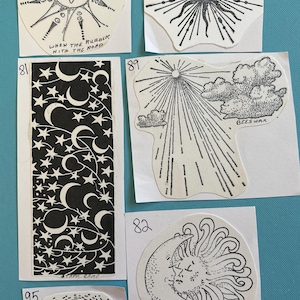 May include: A collection of nine vintage black and white stickers with various celestial designs. The stickers feature images of the sun, moon, stars, and clouds. The stickers are on white paper with handwritten numbers in the top corner of each sticker.