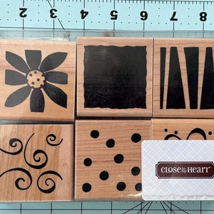 May include: A set of six wooden stamps with different designs. The stamps feature a flower, a swirl, polka dots, a rectangle, and two sets of lines. The stamps are packaged in a clear plastic case with a white card that reads "close to my heart".
