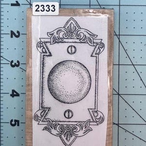 May include: A rectangular wooden stamp with a detailed black and white design. The design features a central dotted sphere within a decorative frame, with the number "2333" on a white label.