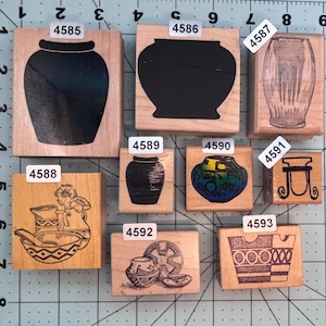 May include: A collection of nine wooden stamps featuring various pottery designs. The stamps include images of vases, bowls, and other ceramic vessels. The designs are in black ink, with some stamps showing intricate patterns and details. The stamps are on a grid background.