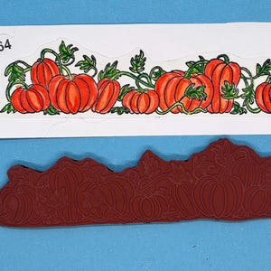 May include: A rubber stamp and a sticker featuring a pumpkin design. The sticker has a white background with a row of orange pumpkins with green vines and leaves. The stamp is a dark red color and has a similar pumpkin design.