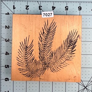 May include: A wooden stamp featuring a black illustration of three stylized feathers. The stamp has a rectangular shape with a light brown surface. The number "7027" is printed on a white label in the upper right corner.