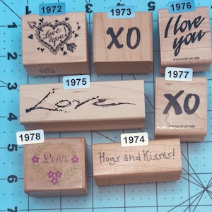 May include: A collection of wooden stamps with various love-themed designs. The stamps feature phrases like "Love You," "XO," and "Hogs and Kisses!" in different fonts and styles. Some stamps include decorative elements like hearts and floral accents.