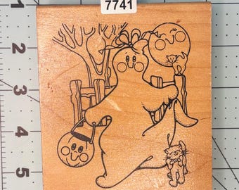 May include: Wooden stamp featuring a Halloween-themed design. The stamp depicts a friendly ghost holding a jack-o'-lantern, with a cat and a smiling moon in the background. The stamp has the number 7741 on it.