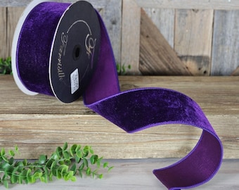 May include: A roll of opulent, deep purple velvet ribbon. The ribbon is unrolled, displaying its plush texture and rich colour. The roll is set against a rustic wooden background, with a sprig of green foliage in the corner.