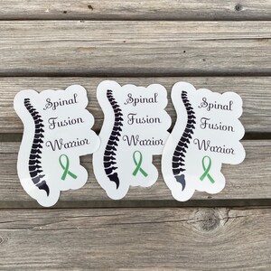 May include: Three white stickers with a black spinal column illustration and green ribbon. The text on each sticker reads "Spinal Fusion Warrior".