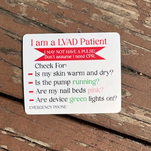 May include: A white card with red text that reads "I am a LVAD Patient." The card also includes instructions for emergency responders, such as checking skin warmth and the pump's function. The card is on a weathered wooden surface.