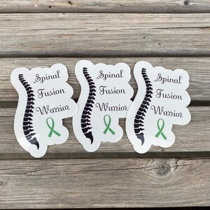 May include: Three white stickers with a black spine illustration and green ribbon. The stickers say "Spinal Fusion Warrior".