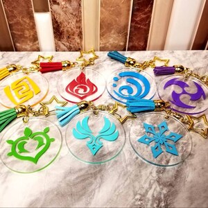 May include: A collection of clear acrylic keychains featuring colorful symbols and tassels. Each keychain has a gold star-shaped charm and a tassel in various colors, including yellow, red, green, blue, and purple. The symbols are in red, yellow, blue, green, and purple.