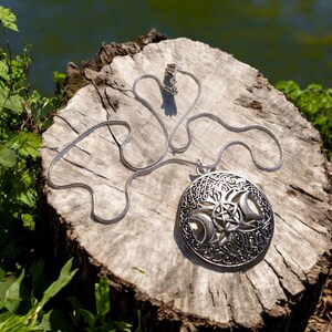 May include: A silver-toned pendant necklace featuring a circular pendant with intricate Celtic knotwork, crescent moons, and a pentagram. The necklace chain is a delicate snake chain. The pendant is displayed on a weathered wooden surface, with greenery in the background.
