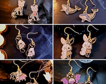 Puede incluir: Una colección de pendientes colgantes dorados con varios diseños temáticos de Halloween. Los pendientes presentan imágenes de gatos, ballenas, conejos y murciélagos en colores negro, blanco, rosa y morado. Los pendientes se muestran sobre una superficie oscura.