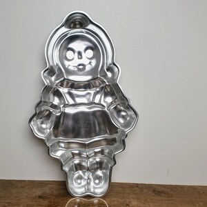 May include: A vintage, silver-colored metal cake pan in the shape of a doll. The pan features a detailed design of a doll with a bonnet, dress, and shoes. The pan is likely used for baking cakes in a unique shape.