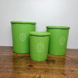 May include: Three green plastic containers with lids. Each container has a white floral design. The containers are different sizes, with the largest on the left and the smallest in the center. The containers are on a wooden surface.