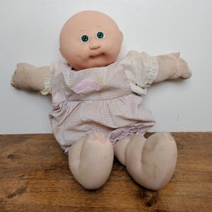 May include: A bald doll with a round face, green eyes, and dimpled cheeks. The doll wears a light pink and white chequered jumper with a small pink embroidered logo on the chest over a white short-sleeved shirt with ruffled sleeve edges.