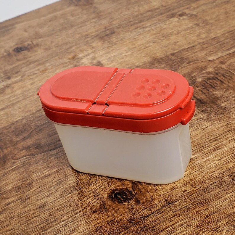 Pu&ograve; includere: Un contenitore per spezie in plastica bianca con un coperchio rosso. Il coperchio ha un'apertura a cerniera con un piccolo motivo a buchi per dispensare le spezie.