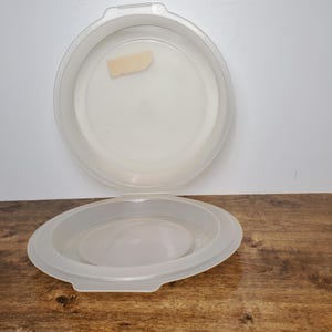 May include: A clear plastic cake container with a lid. The container is round and has a smooth surface. The lid has a small label on it.