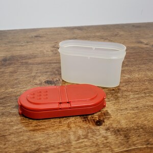 Pu&ograve; includere: Un contenitore per spezie in plastica rossa con un coperchio in plastica trasparente. Il coperchio ha un piccolo foro al centro per dispensare le spezie.