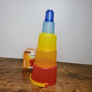May include: A set of four colorful plastic stacking cups, with a blue, light blue, yellow, orange, and red color scheme. The cups are stacked on top of each other, with the largest cup at the bottom and the smallest cup at the top. The cups are made by Tupperware.