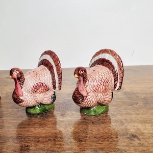 May include: A set of two ceramic turkey salt and pepper shakers. The turkeys are brown, white, and pink with detailed feathers. They are standing on a green base.