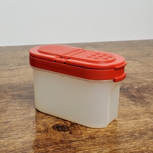 Pu&ograve; includere: Un contenitore per spezie in plastica trasparente con un coperchio rosso. Il coperchio ha una parte superiore incernierata e una fila di fori per spolverare.
