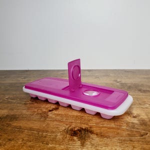 May include: A magenta and white ice cube tray with a built-in stand. The tray has a rectangular shape with rounded edges and a series of individual ice cube molds. The stand is attached to the top of the tray.