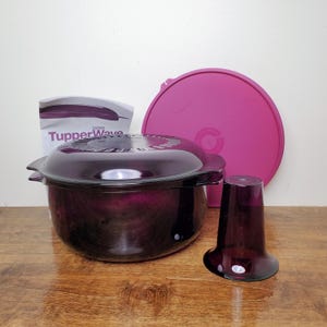 May include: A set of Tupperware microwave cookware in a deep purple hue. The set includes a round casserole dish with a lid, a separate pink lid, and a small, flared glass. A package with the Tupperware logo is also present.