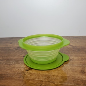 May include: Collapsible green and white silicone bowl with a matching green lid. The bowl is open and expanded, showing the accordion-like design. The lid sits beneath the bowl on a wooden surface.