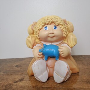 May include: A vintage Cabbage Patch Kids doll figurine with blonde hair and blue eyes, holding a blue piggy bank. The doll is wearing a peach-coloured dress and has white shoes. A nostalgic collectible.