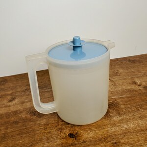May include: A clear plastic pitcher with a blue lid and a handle. The pitcher is on a wooden surface.