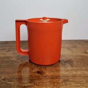 May include: A bright orange plastic pitcher with a white lid and a small knob on top. The pitcher is sitting on a wooden surface.
