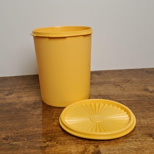 May include: A yellow plastic container with a lid. The container is tall and cylindrical. The lid has a sunburst design.